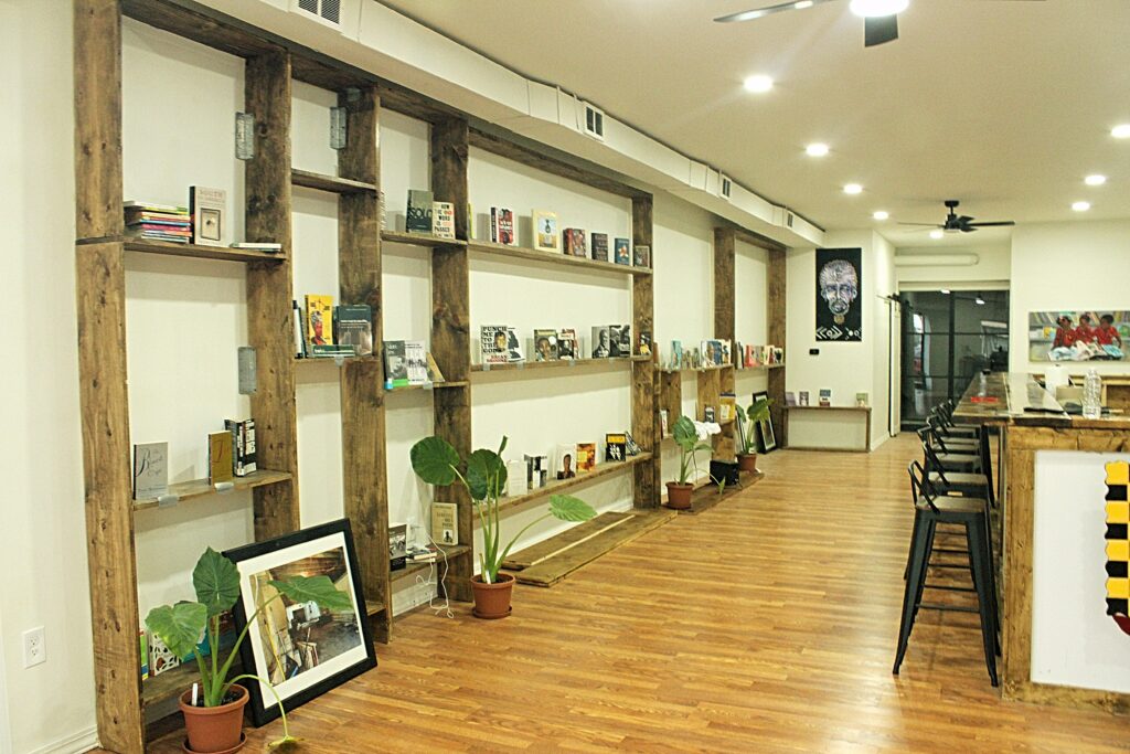 Jerjuan Howard: Community Enhancement Through Literacy Image 8 Sparcely spaced rustic wooden shelves on a white wall, featuring several books. Cafe counter with metal stools.