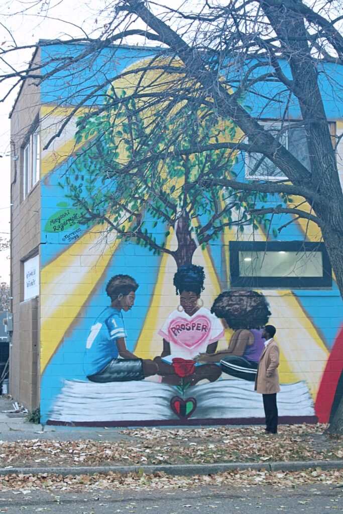 Jerjuan Howard: Community Enhancement Through Literacy Image 9 Mural of 3 black children sitting on an opened book under a tree, with the word "Prosper" inside a pink heart floating in front of them.