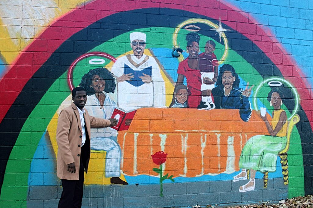 Jerjuan Howard: Community Enhancement Through Literacy Image 3 Jerjuan Howard, a dapper young black man, standing next to a multicolored mural of an orange table with six seated black figures of various ages, many with halos.