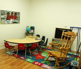 Children's reading corner at Howard Family Bookstore in Detroit. Photo ©2025 by Ryan M Place. All rights reserved. Arc-shaped table with 6 small, child-sized chairs on a USA Map/Alphabet area rung. Adult sized wooden rocking chair, and a painting of 3 black children reading books.