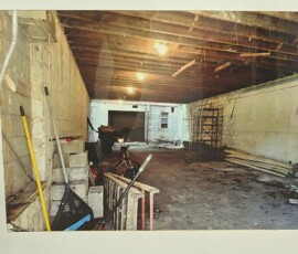 Basement of Howard Family Bookstore in Detroit. Photo ©2025 by Ryan M Place. All rights reserved. Basement space with dingy white cinderblock walls and a dusty concrete floor, wooden floor joist forming the ceiling. Metal shelves and several construction, yard, and cleaning implements are visible.