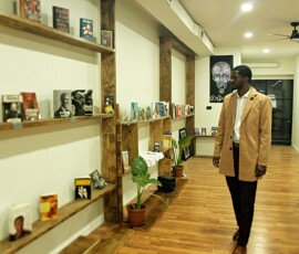 Jerjuan Howard inside Howard Family Bookstore in Detroit. Photo ©2025 by Ryan M Place. All rights reserved. Jerjuan Howard, a dapper young black man, in black pants and a tan overcoat looking at rustic bookshelves.