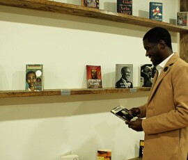 Jerjuan Howard inside Howard Family Bookstore in Detroit. Photo ©2025 by Ryan M Place. All rights reserved. Profile photo of Jerjuan Howard, a black man, holding a Malcolm X book in front of rustic wooden bookshelves set against a white wall.