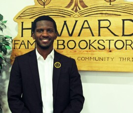Jerjuan Howard, interior of Howard Family Bookstore in Detroit. Photo ©2025 by Ryan M Place. All rights reserved. Handsome black man with a moustache standing in front of a handcrafted wooden sign reading "HOWARD FAMILY BOOKSTORE".