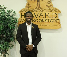 Jerjuan Howard at Howard Family Bookstore in Detroit. Photo ©2025 by Ryan M Place. All rights reserved. Jerjuan Howard, a dapper young black man in a black suit, standing in front of a handcrafted wooden sign on a white wall.