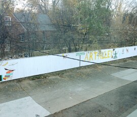 Puritan Avenue Art Alley. Photo ©2025 by Ryan M Place. All rights reserved. Looking down across the street from a white-painted picket fence reading "Puritan Ave ART ALLEY".
