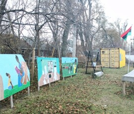 Umoja Community Garden in Detroit. Photo ©2025 by Ryan M Place. All rights reserved. Grassy field with fallen leaves, featuring 3 murals on billboards set against leafless trees. A yellow-painted shipping container, metal shelves, and a rustic wooden table are also featured.