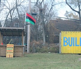 Umoja Community Garden in Detroit. Photo ©2025 by Ryan M Place. All rights reserved. Fruit/vendor stand with sign reading "Many Hands Make Light Work" and a yellow shipping container with blue text reading "BUILD" in a grassy field.