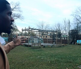 Jerjuan Howard talking about Umoja Community Garden in Detroit. Photo ©2025 by Ryan M Place. All rights reserved. Jerjuan Howard, a young black ban, in a grassy field featuring a large wooden structure and some murals painted on billboards.