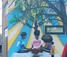 Exterior Mural at Howard Family Bookstore in Detroit. Photo ©2025 by Ryan M Place. All rights reserved. Mural of 3 black children sitting on an opened book under a tree, with the word "Prosper" inside a pink heart floating in front of them.