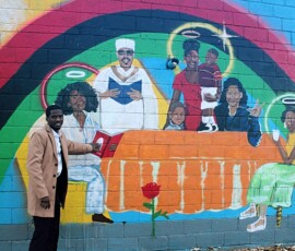 Jerjuan Howard in front of mural at Howard Family Bookstore in Detroit. Photo ©2025 by Ryan M Place. All rights reserved. Jerjuan Howard, a dapper young black man, standing next to a multicolored mural of an orange table with six seated black figures of various ages, many with halos.